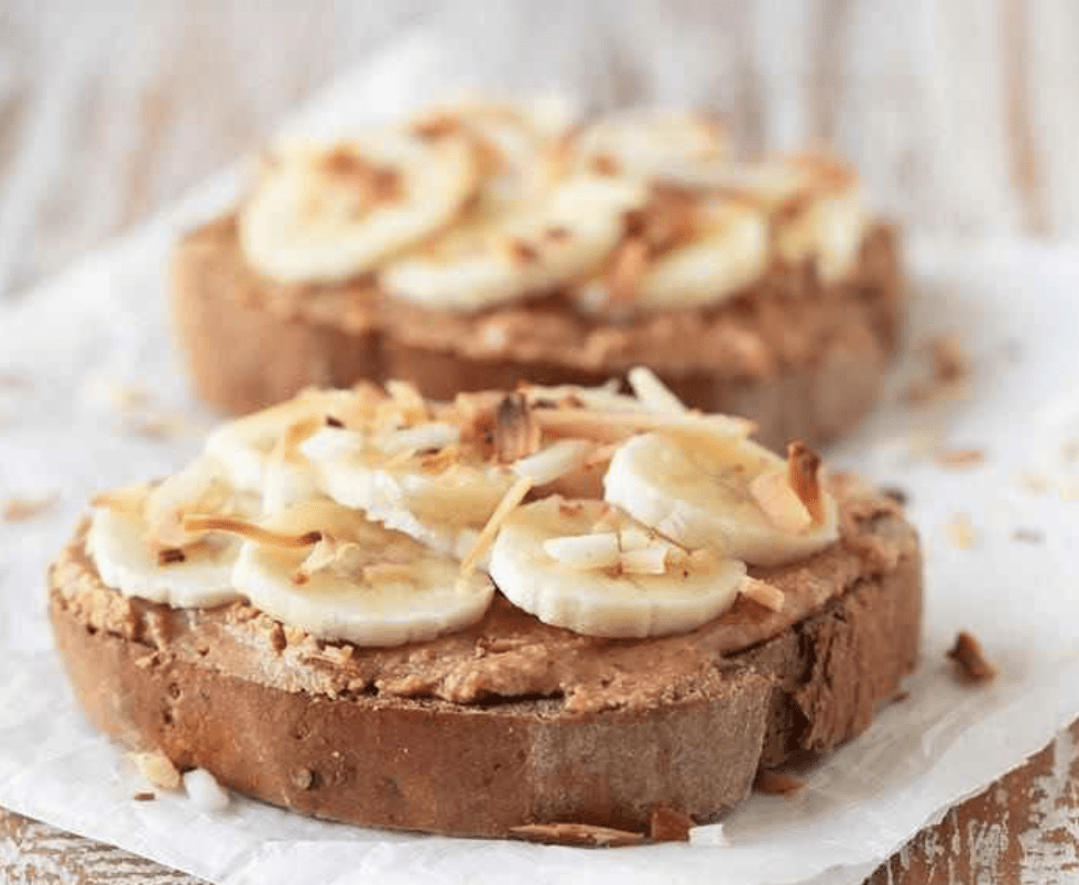 Tostadas integrales con crema de cacahuate, rodajas de plátano y coco tostado, servidas sobre papel pergamino.